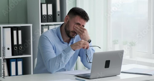 Caucasian businessman sits at his desk, struggling to keep his eyes open. He removes glasses, massages temples, and takes a deep breath in an effort to push through his exhaustion.