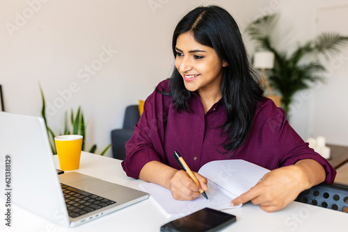 Canvas Print Young adult indian student woman taking notes while using laptop computer at home