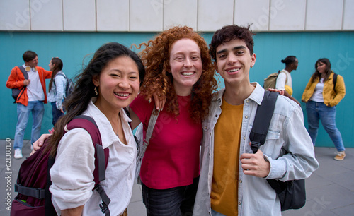 Foto Group portrait of three smiling multiracial students hugging backpacks looking at camera standing outside college campus