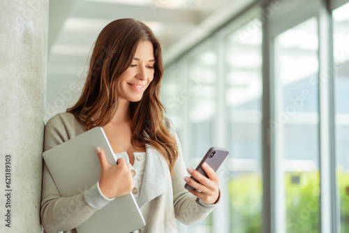 Woman with beautiful hair wearing casual clothes holding mobile phone checking mail in modern office