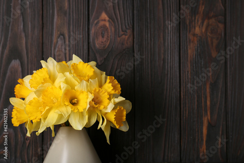 Bouquet of beautiful yellow daffodils in vase near wooden wall. Space for text