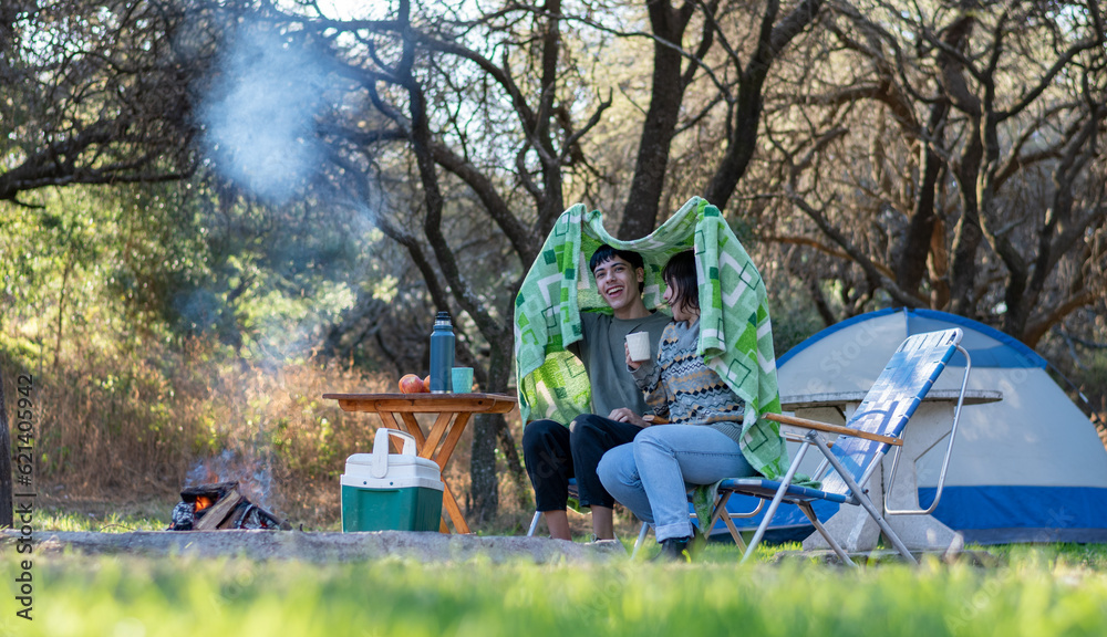 Two funny and cold friends drinking tea covered with a blanket. Campfire at the campsite Two funny and cold friends drinking tea covered with a blanket. Campfire at the campsite