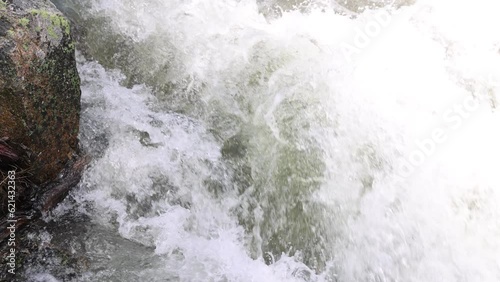 Green and white creek water rushes forcefully past rocks on the bank. Slow motion photo