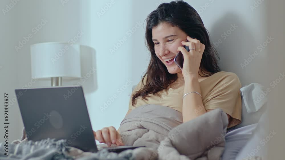 Young brunette girl sitting in bed, talking on mobile phone and using laptop in the morning at home. Medium shot Young brunette girl sitting in bed, talking on mobile phone and using laptop in the morning at home. Medium shot