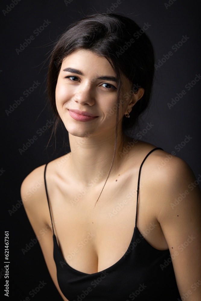 Classic studio portrait of a young brunette dressed in a black top, who is sitting on a chair against a black background.