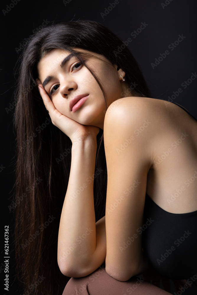 Classic studio portrait of a young brunette dressed in a black top, who is sitting on a chair against a black background. Classic studio portrait of a young brunette dressed in a black top, who is sitting on a chair against a black background.