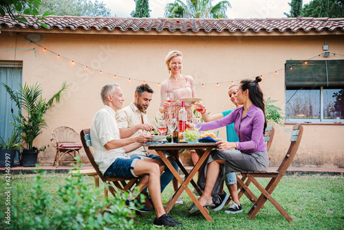 A group of mature adult friends having fun together on a barbeque lunch party at home backyard. Middle aged happy people smiling celebrating a birthday dining reunion with family at patio outside