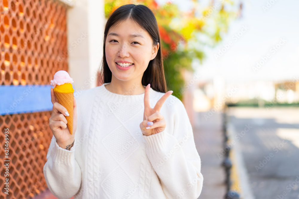 Pretty Chinese woman with a cornet ice cream at outdoors smiling and showing victory sign Pretty Chinese woman with a cornet ice cream at outdoors smiling and showing victory sign