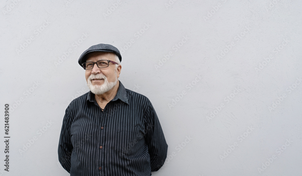 An elderly smiling man in glasses and a cap stands against a light wall background, hands behind his back, horizontal format An elderly smiling man in glasses and a cap stands against a light wall background, hands behind his back, horizontal format