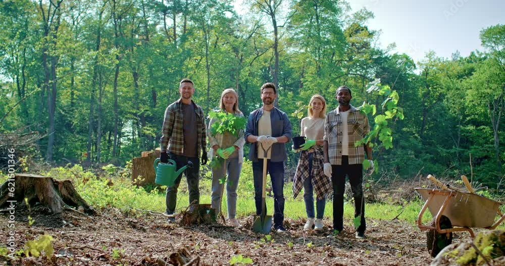 Joyful ecological activists men and women are holding garden tools and plants, standing on background of forest looking at camera. Volunteering, charity work and lifestyle concept. Joyful ecological activists men and women are holding garden tools and plants, standing on background of forest looking at camera. Volunteering, charity work and lifestyle concept.