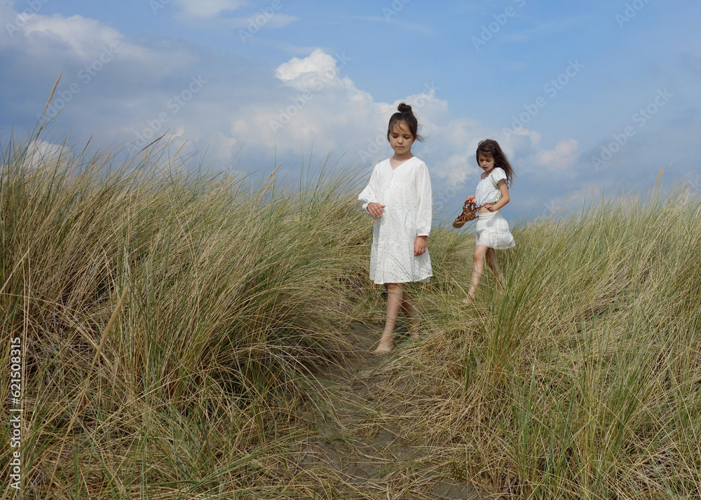 Two girls in white dresses walking between sand grass on dunes Two girls in white dresses walking between sand grass on dunes