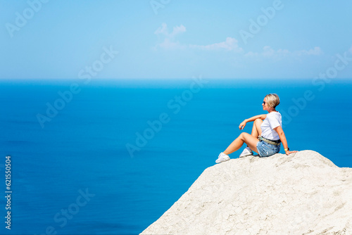 A young woman in denim shorts and a white T-shirt sits on a cliff against the backdrop of the sea and blue sky on a sunny day. Active recreation, travel and tourism.