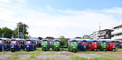 BANGKOK, THAILAND - JULY 10, 2023: Several Electric tuk tuks taxi stopped in the meadow with buildings background. Concept to save fuel, maintain and reduce air pollution protect the environment.