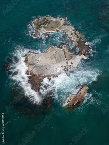 A wreaked ship resting on a rock half submerged from a drone view