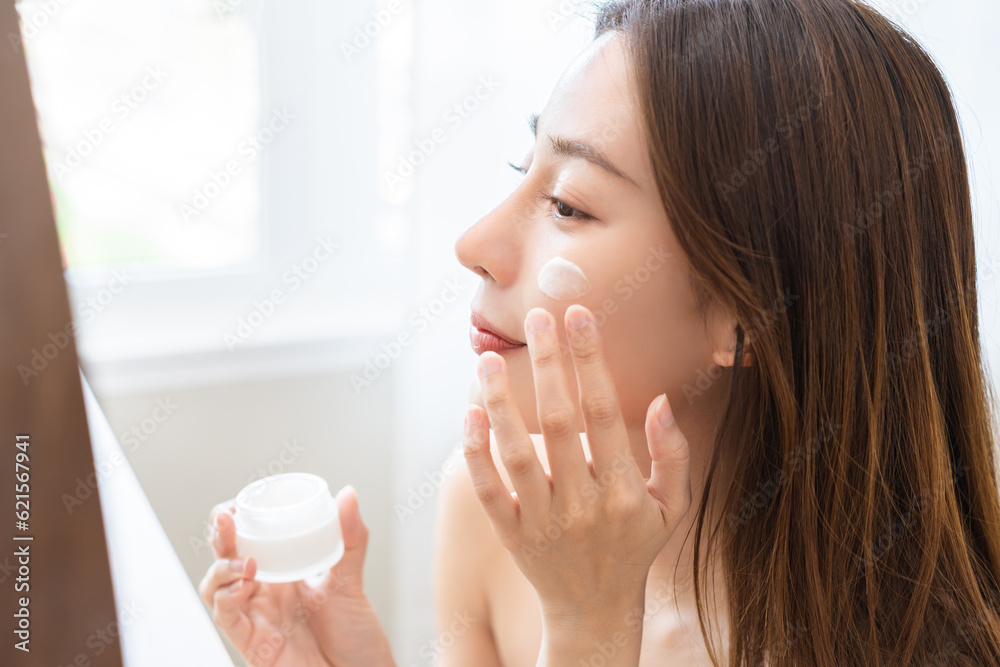 Facial beauty skin care, smile of pretty asian young woman in bathrobe looking at mirror, hand applying moisturizer lotion on her face, holding jar of skin cream before makeup cosmetic routine at home Facial beauty skin care, smile of pretty asian young woman in bathrobe looking at mirror, hand applying moisturizer lotion on her face, holding jar of skin cream before makeup cosmetic routine at home