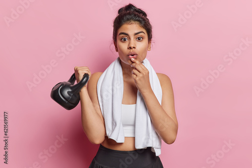 Horizontal shot of scared young sporty woman with hair bun lifts weight poses in sportswear with white towel around neck reacts to something shocking isolated over pink background. Sport and lifestyle
