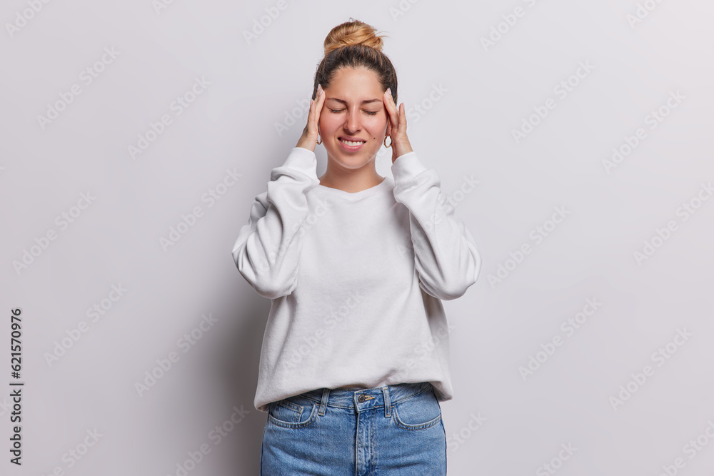 Photo of young woman suffers from headache keeps hands on temples keeps eyes closed dressed in casual jumper and jeans feels unbearable migraine poses against white background cannot concentrate Photo of young woman suffers from headache keeps hands on temples keeps eyes closed dressed in casual jumper and jeans feels unbearable migraine poses against white background cannot concentrate