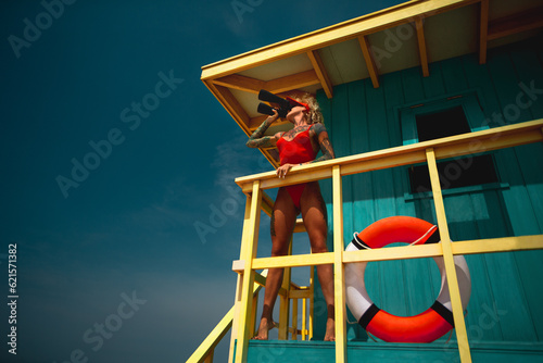 Lifeguard woman in red bikini and visor with binocular and lifebuoy on watch tower against turquoise sky