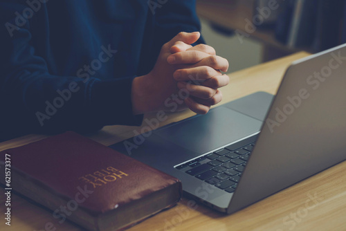 Man's hands are folded in prayer on a Holy Bible over a laptop in church concept for faith, religion, love, and forgiveness. Holy Bible study reading together in Sunday school.Online church