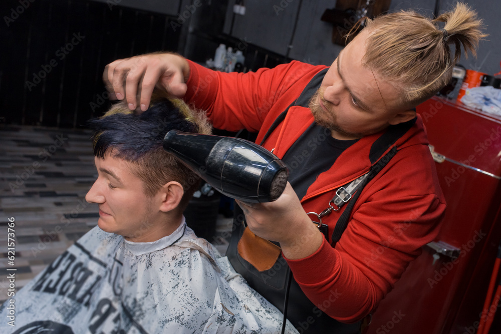 Naklejka premium A stylish adult barber European who is passionate about work dries his hair with a hair dryer for a young guy, a Caucasian client in a salon