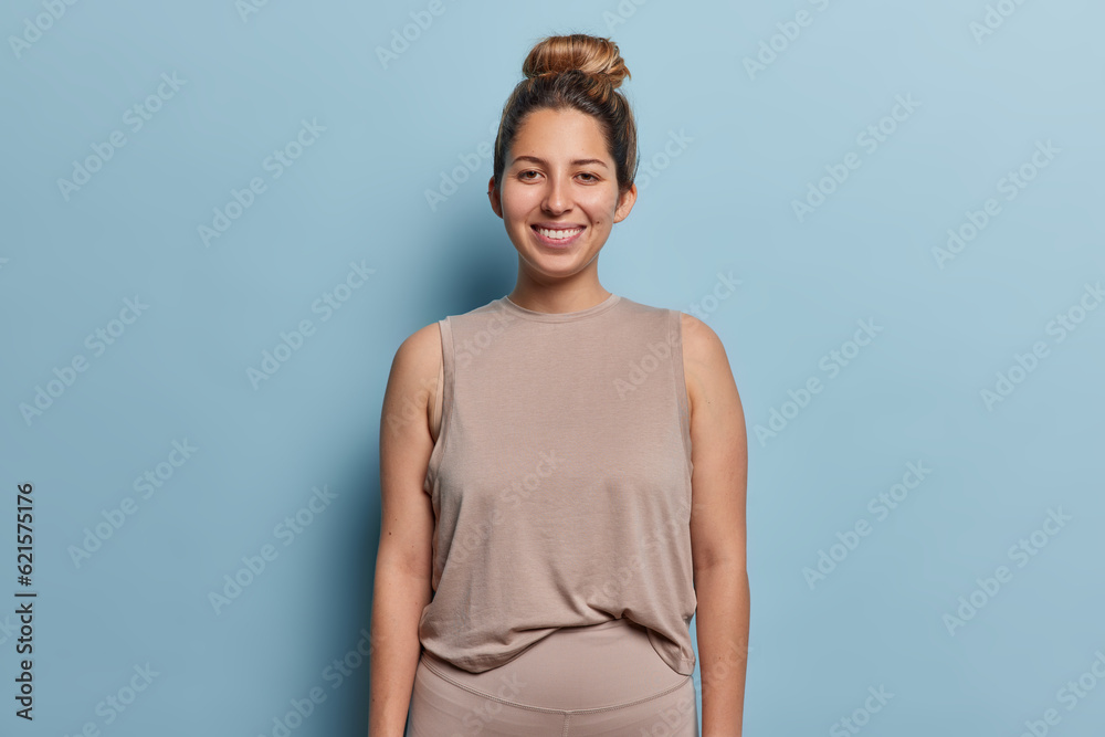 Studio shot of positive young woman with hair bun smiles gladfully dressed in beige tshirt and leggings being in good mood after training in gym isolated over blue background. Sporty lifestyle concept Studio shot of positive young woman with hair bun smiles gladfully dressed in beige tshirt and leggings being in good mood after training in gym isolated over blue background. Sporty lifestyle concept