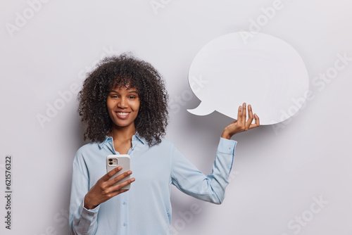 Horizontal shot of pleased curly haired young woman holds modern smartphone and speech bubble for your promotional content wears formal clothes being in good mood isolated over white background
