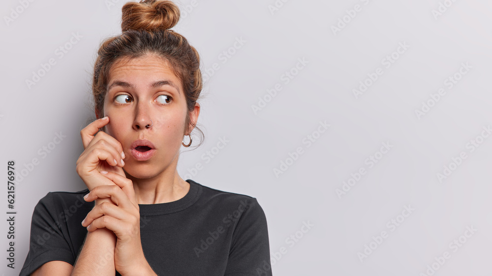 Horizontal shot of shocked young woman with hair gathered in bun looks stunned aside holds breath from amazement wears casual black t shirt isolated on white background with copy space for your promo Horizontal shot of shocked young woman with hair gathered in bun looks stunned aside holds breath from amazement wears casual black t shirt isolated on white background with copy space for your promo