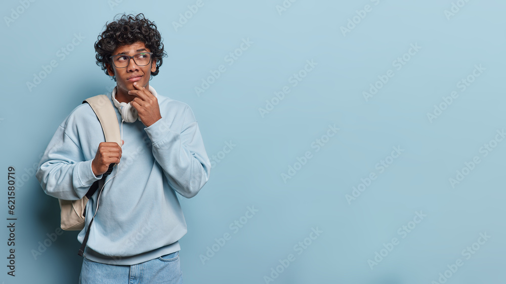 Studio shot of isolated on left side of blue background young thoughtful hindu man with short black curly hair wearing blue jeans and sweater with white big bag on left shoulder and headphones on neck Studio shot of isolated on left side of blue background young thoughtful hindu man with short black curly hair wearing blue jeans and sweater with white big bag on left shoulder and headphones on neck