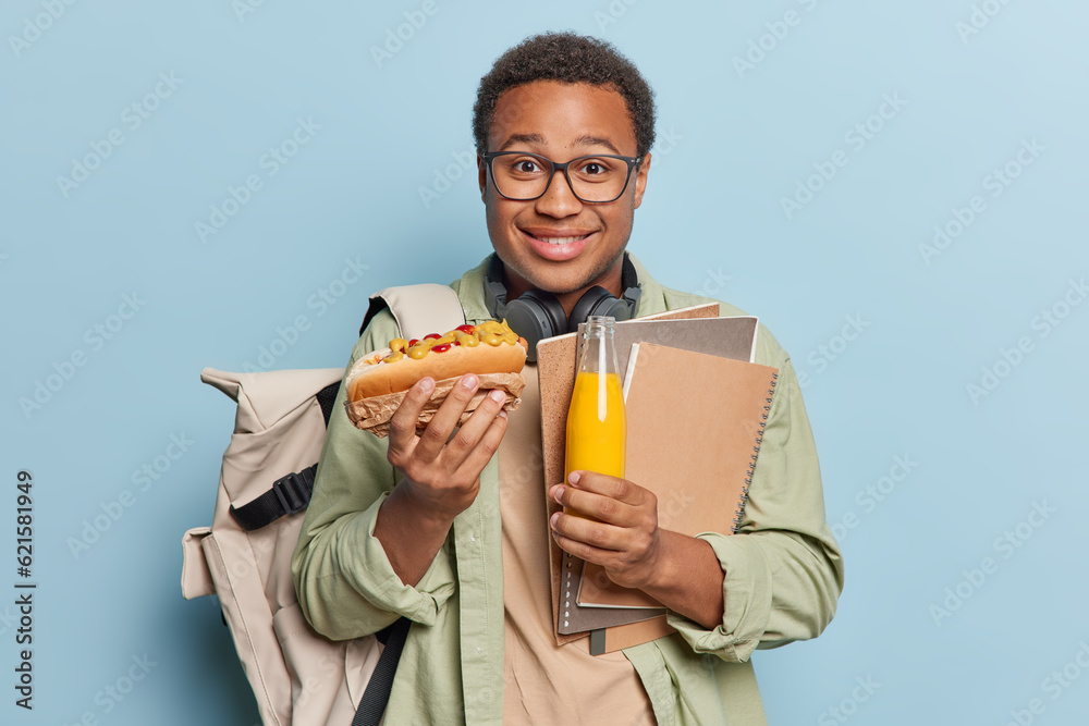 Indoor shot of positive dark skinned man eats hot dog holds bottle of fresh juice carries notepads carries rucksack smiles pleasantly dressed in casual clothes isolated over blue background. Indoor shot of positive dark skinned man eats hot dog holds bottle of fresh juice carries notepads carries rucksack smiles pleasantly dressed in casual clothes isolated over blue background.