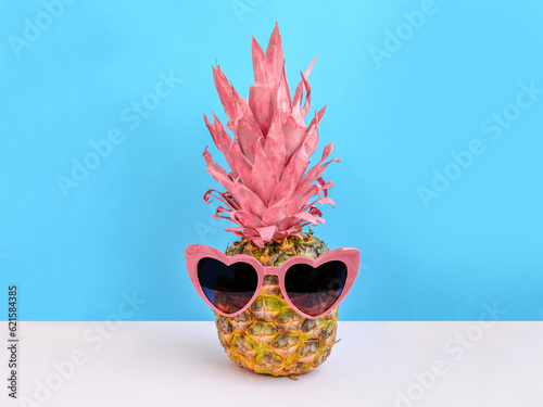 Creative summer layout made of pineapple with pink leaves and heart shaped sunglasses against blue and pink background. Original pineapple decoration. Creative summer idea. Fruit concept. Copy space.
