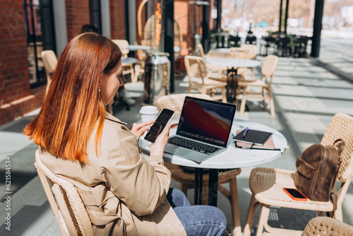 Young woman sitting at table and using smartphone and laptop outdoors. Online education, order, working or shopping concept