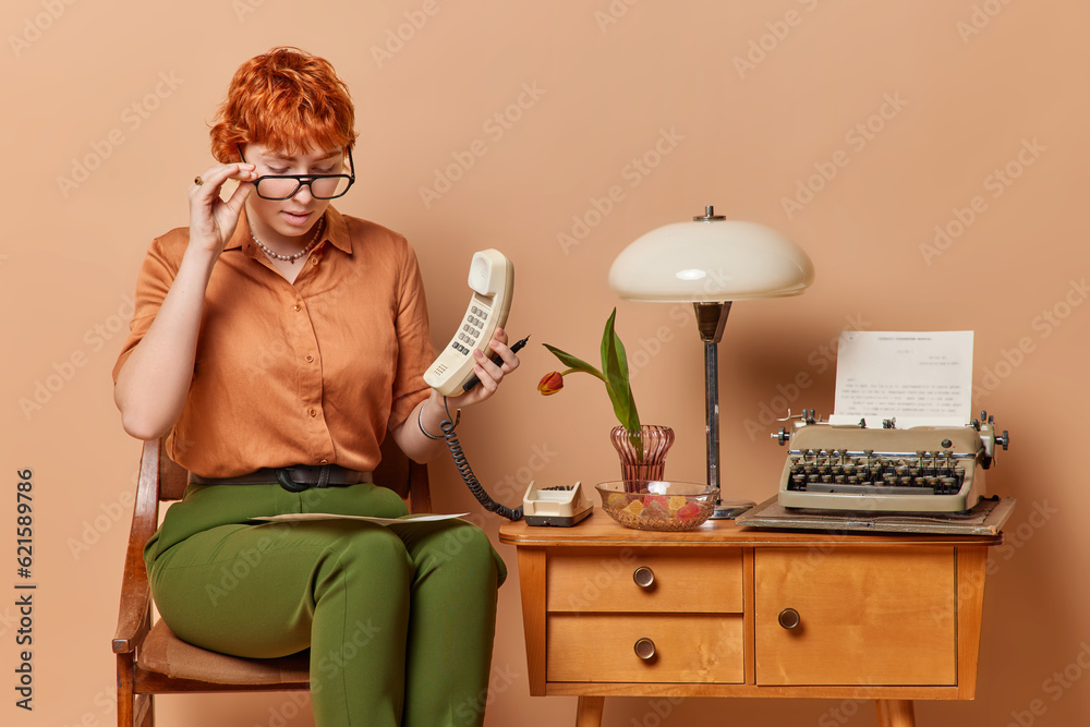 Attentive busy redhead young businesswoman or secretary wears spectacles concentrated at paper document holds handset studies financial report isolated over brown background. Retro style concept