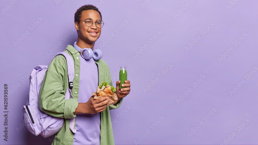 Horizontal shot of cheerful dark skinned guy eats junk food and drinks healthy green smoothie carries rucksack wears green shirt headphones around neck isolated over purple background copy space