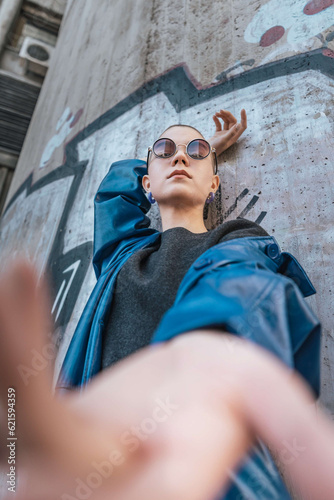 Wide angle portrait of a short hair young female wearing circular sunglasses and blue coat leaning on a concrete wall.