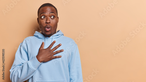 Horizontal shot of dark skinned bearded man feels shocked keeps hand on chest has bugged eyes holds breath from amazement wears casual blue sweatshirt isolated over brown background. Omg concept