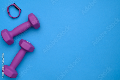 The layout of two rubberized dumbbells of 2 kg of purple color, a fitness bracelet on a blue background, top view.