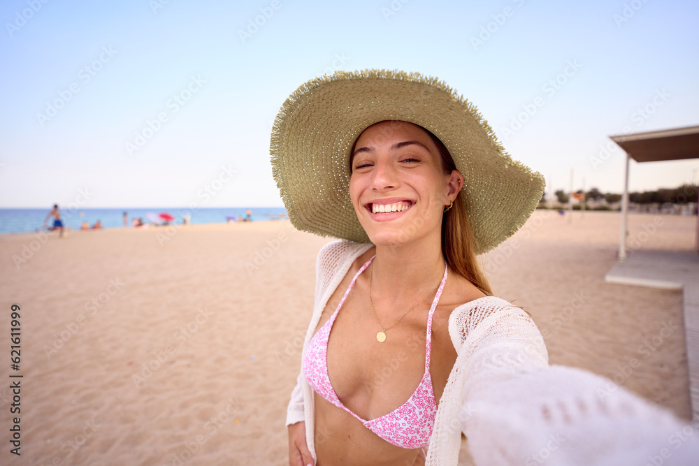 Portrait of young Caucasian woman in her twenties wearing straw hat smiling and taking selfie while walking on beach. Attractive gen z blonde girl enjoying summer vacation by Mediterranean outdoors.