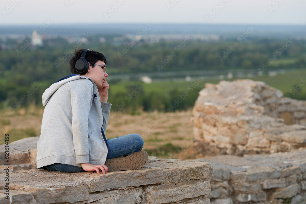 A teenage boy with headphones listening to music on the ruins of an ancient fortress. In the background is a blurry image of the cityscape.