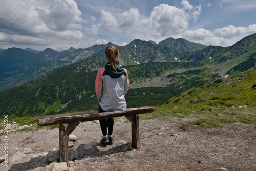 Young girl is sitting on the bench enjoying view of high peaks of Vysoke Tatry mountains near to Zuberec town. Bench located on Spalena dolina near to mountain lakes and chairlift station. Slovakia.