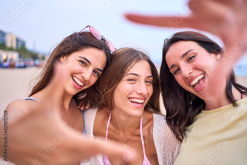 Happy group of three beautiful Caucasian friends taking selfie doing hand frame gesture on beach. Smiling girls looking through fingers at camera. Young women having fun outdoors on summer vacation. photo