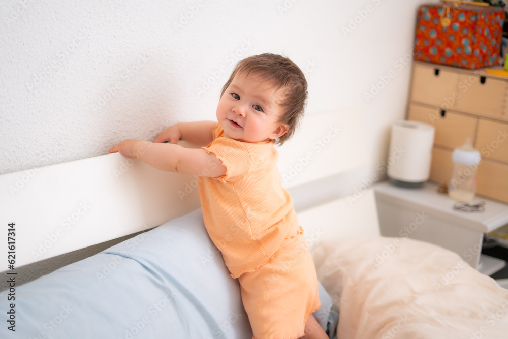 lifestyle home portrait of adorable and beautiful 9 months old baby girl learning to stand up by holding bed frame smiling cheerful and proud in childhood concept lifestyle home portrait of adorable and beautiful 9 months old baby girl learning to stand up by holding bed frame smiling cheerful and proud in childhood concept