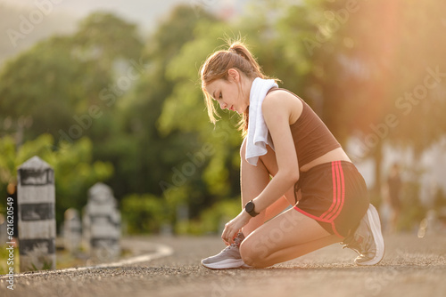 Female runner kneeling and tying shoelace on the street. Fitness woman tying shoelaces. Young woman runner tying shoelaces in park with sunset and sunbeam. © Treerat