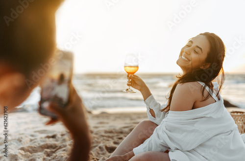 Woman taking a photo of her happy girlfriend at the beach, having picnic and drinking wine at sunset on coasline photo