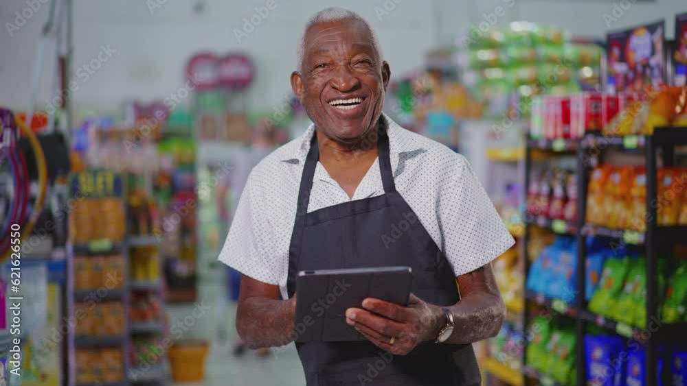 Joyful Happy Brazilian senior employee of supermarket wearing apron and holding tablet device, portrait of a black older male staff depicting job occupation at retail store Joyful Happy Brazilian senior employee of supermarket wearing apron and holding tablet device, portrait of a black older male staff depicting job occupation at retail store