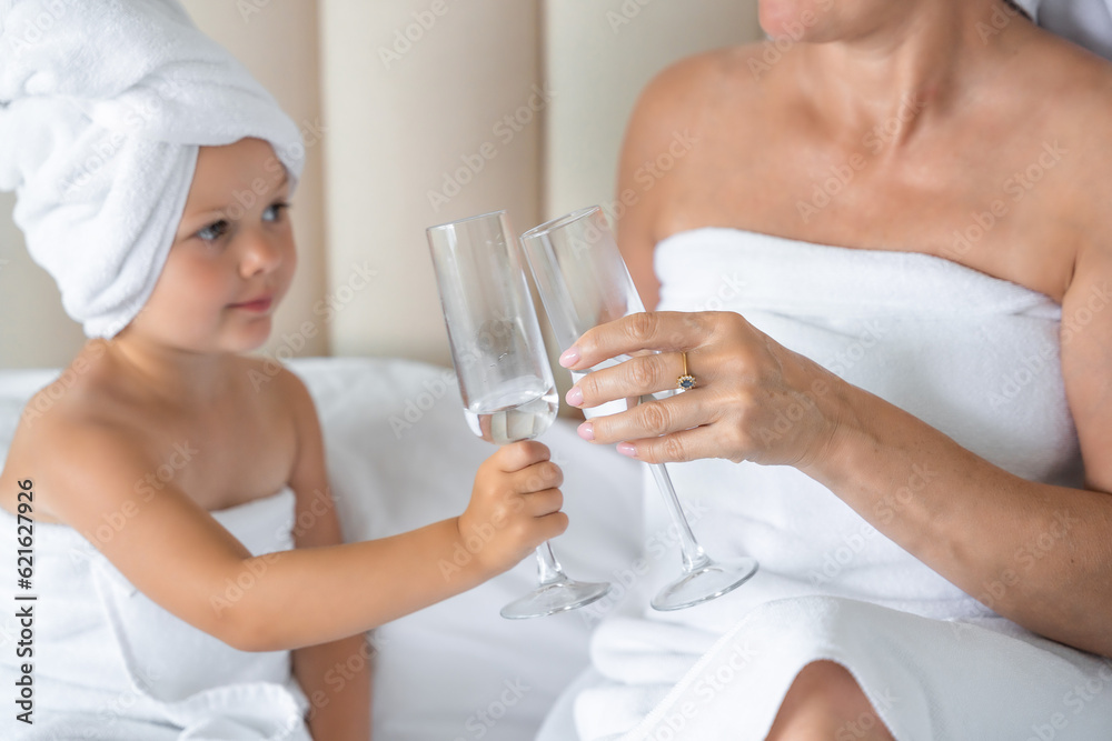 Happy family mother and little daughter in dressing gowns and towels laugh and drinking water with glasses during spa day at home