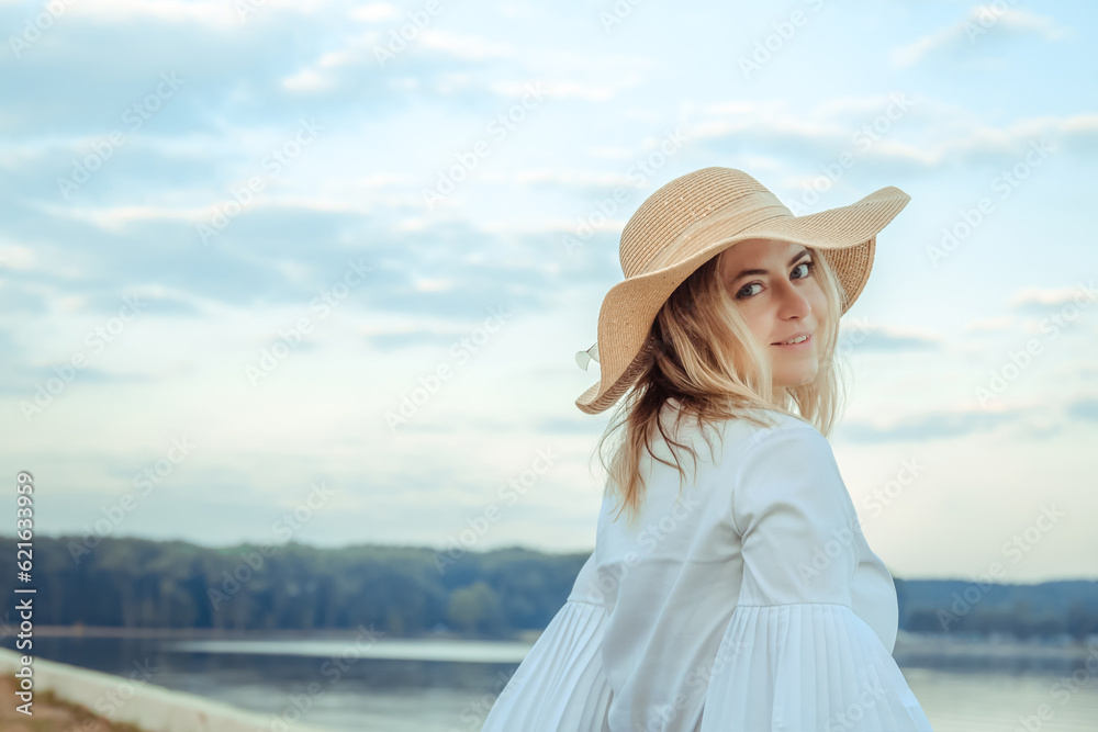 Portrait of young 30 year smiling european woman in straw hat and white dress looking at camera on a cloudy sky background. Beautiful girl. Side view. Profile. Romance mood. International woman day