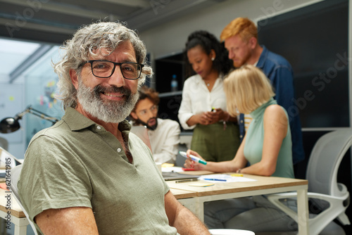 Portrait of an office man smiling and looking at camera. In the background there is a group of employees kipping at work. It is an indoor scene. photo
