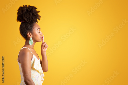 Smiling calm millennial african american lady in dress making shh gesture, putting finger to lips