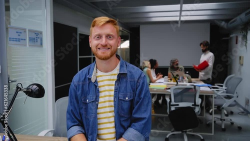 Portrait of an office smiling young man and looking at camera. In the background there is a group of employees kipping at work. It is an indoor scene. photo