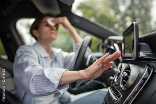 Woman drives car with broken air conditioning in hot summer weather wipes sweat on forehead with paper napkin. Middle aged female suffering from heat stuffiness. Exhausted overheated stressed driver. photo